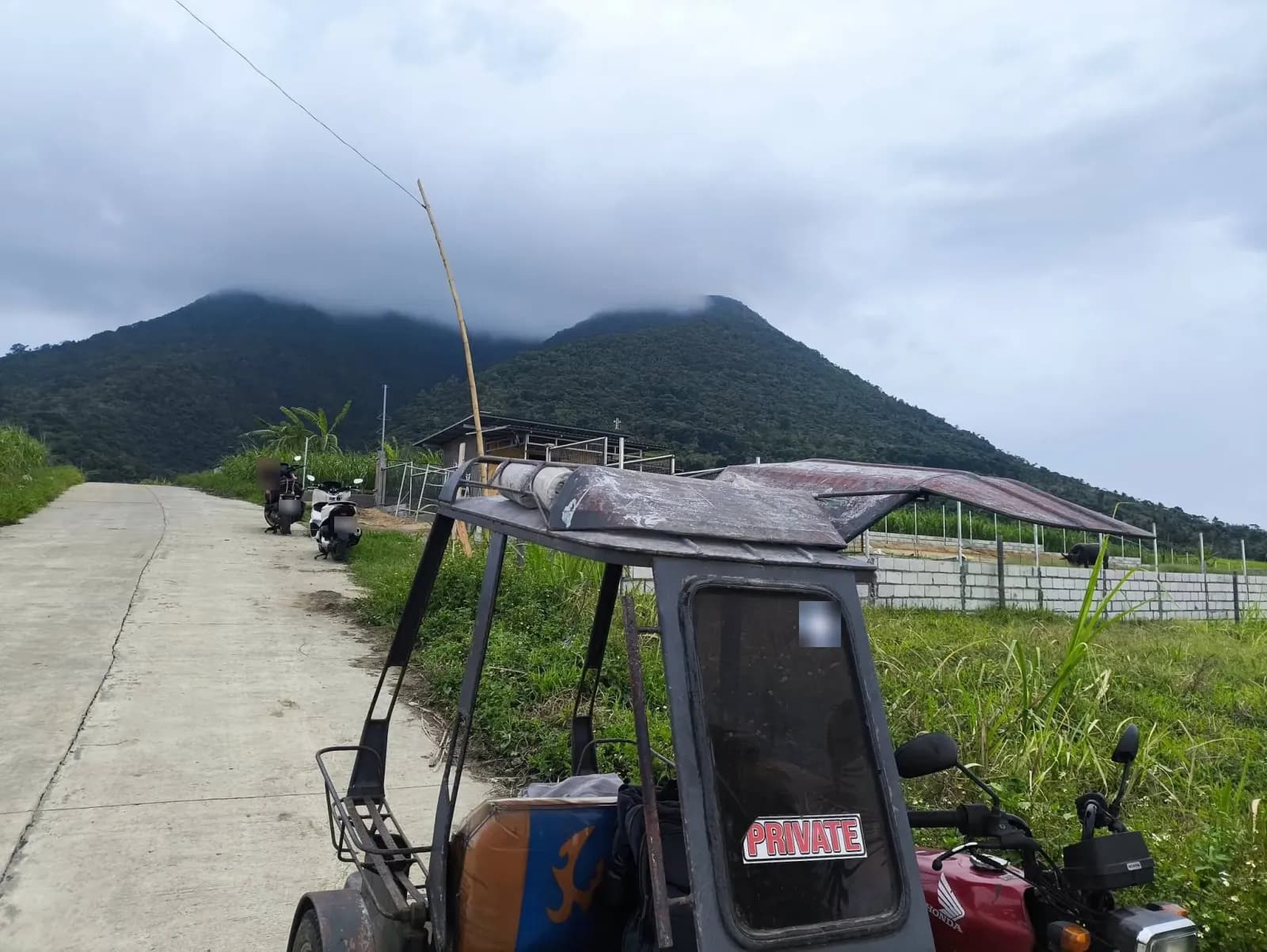 A concrete road beside parked motorcycles and a tricycle, with a low wall, a roofed structure, and a wooded mountain ridge under low cloud