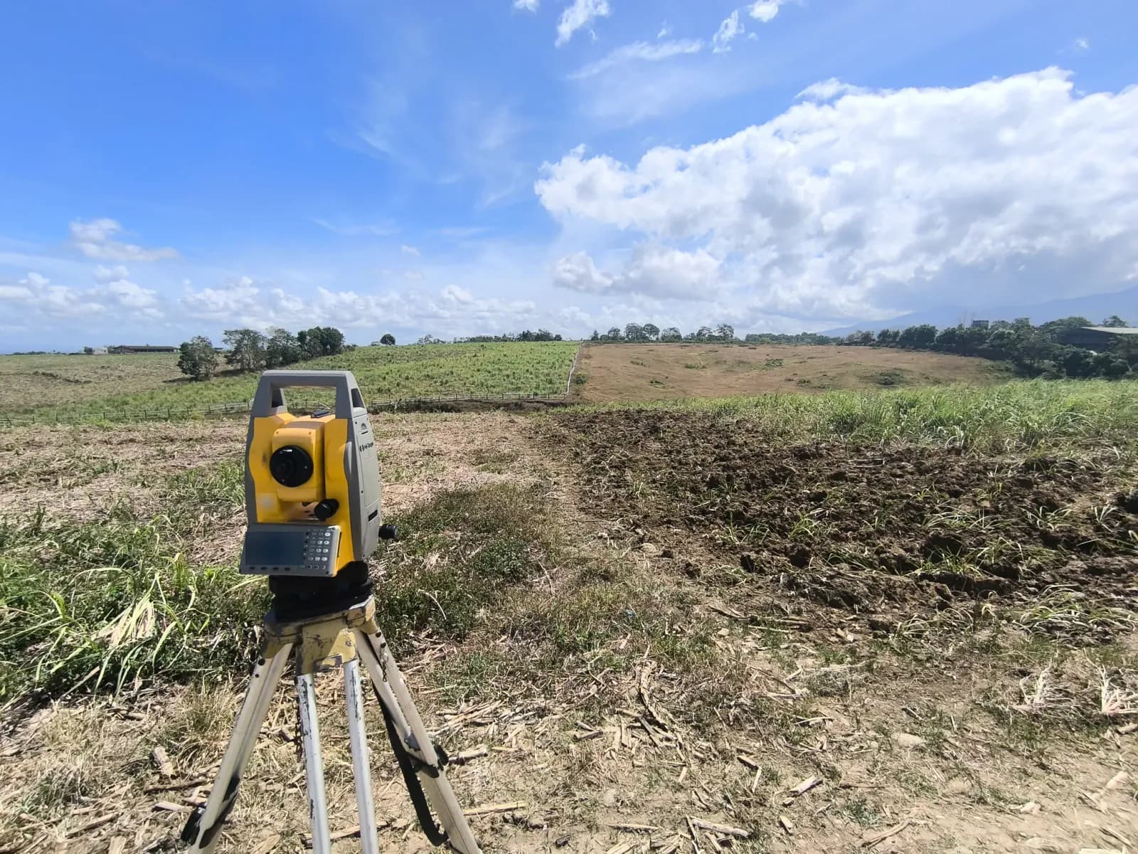 Yellow total station on a tripod beside tilled soil and cropped fields, with a low fence, distant trees, and large clouds above