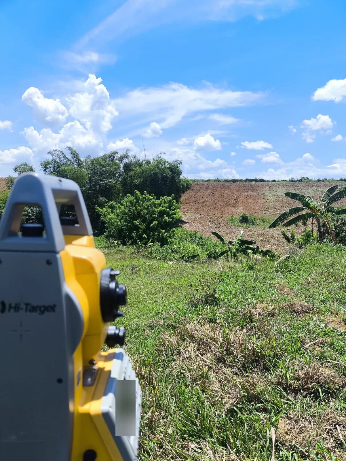 Close view of a total station overlooking a grassy dip, banana plants, bamboo, and a broad patch of exposed soil on the opposite slope
