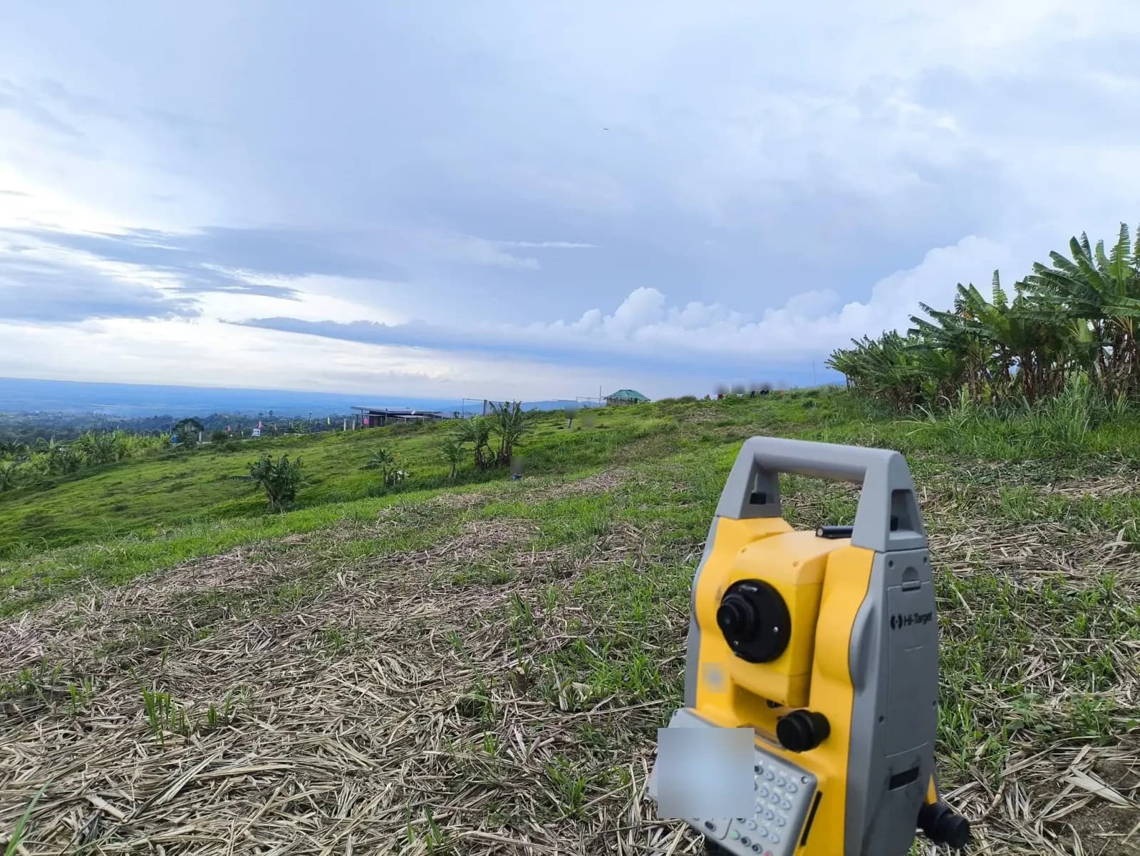 Total station in the foreground overlooking a grassy hillside, banana plants, and people farther across the site beneath a cloudy sky