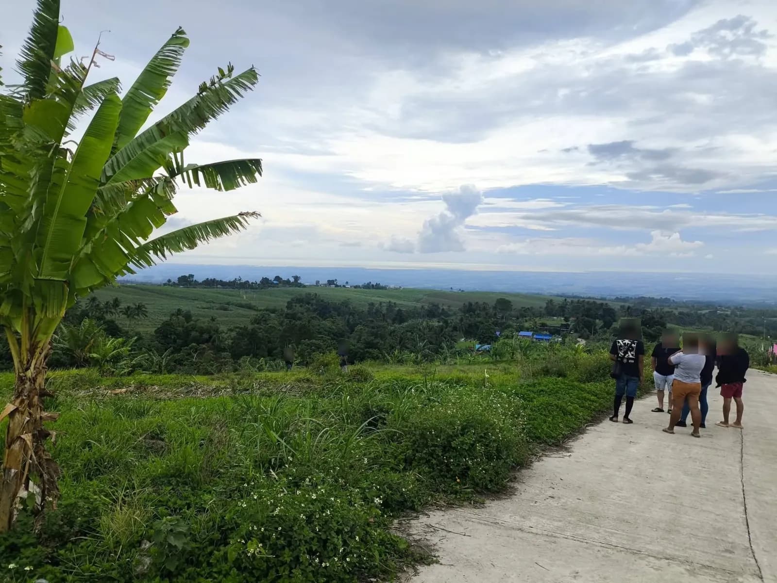 A small group talking beside a hillside road, with banana plants in the foreground and views across green fields, trees, and scattered buildings