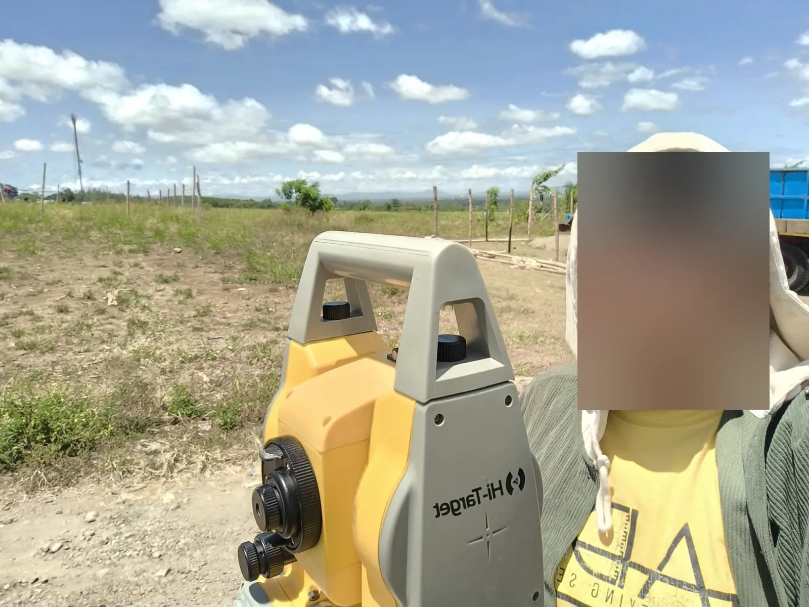 A person beside a yellow total station in an open field, with fence posts, patchy grass, a blue truck, and a bright cloudy sky behind them
