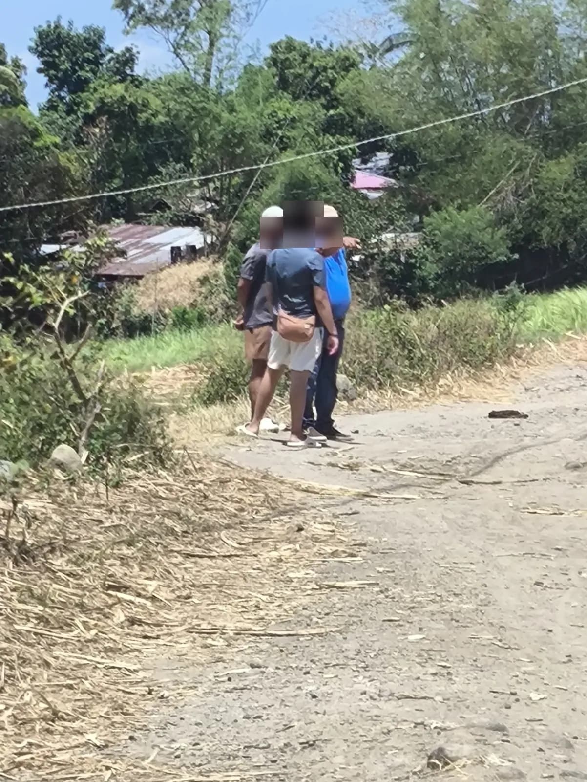 Three people discussing the site beside a narrow road, with one pointing toward the fields and trees and small metal-roofed buildings behind them