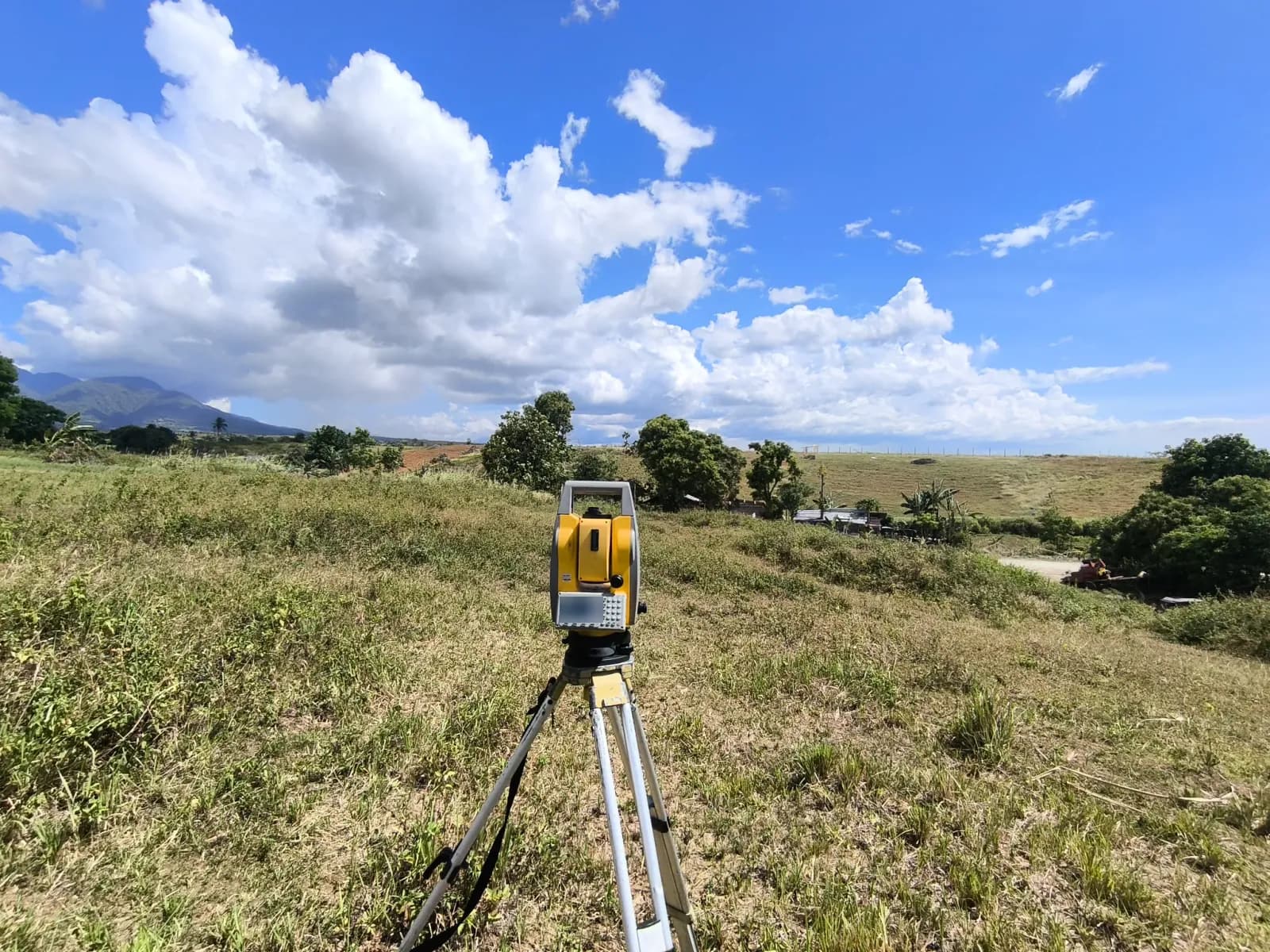 Tripod-mounted total station in an open grassy field, with trees, a mountain ridge, and large white clouds in the distance