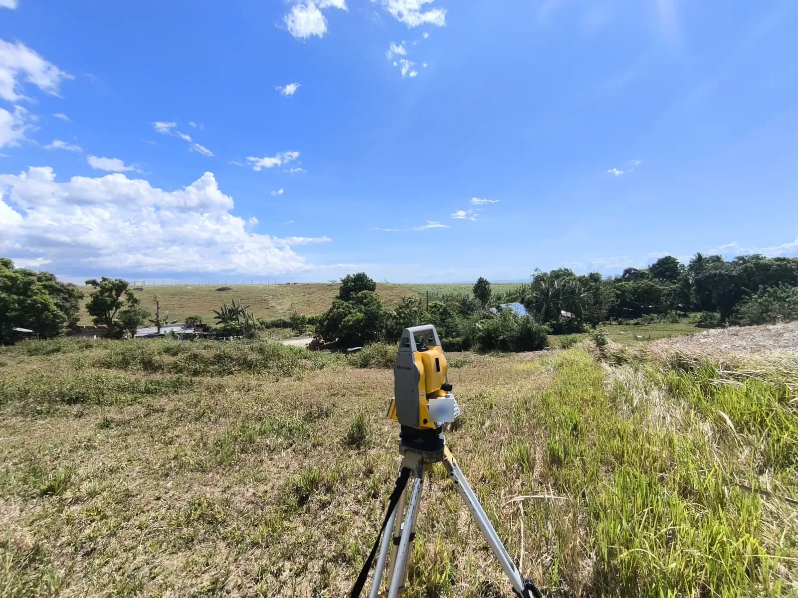 An angled view of a tripod-mounted total station overlooking grassy fields, nearby roofs, and trees, with tall grass along the right edge