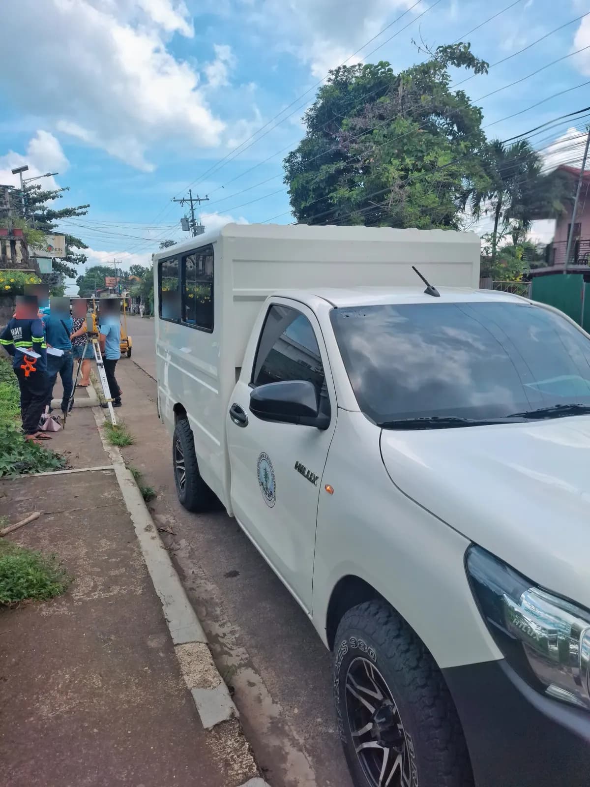 Survey crew gathered beside a total station on the sidewalk next to a parked white utility vehicle