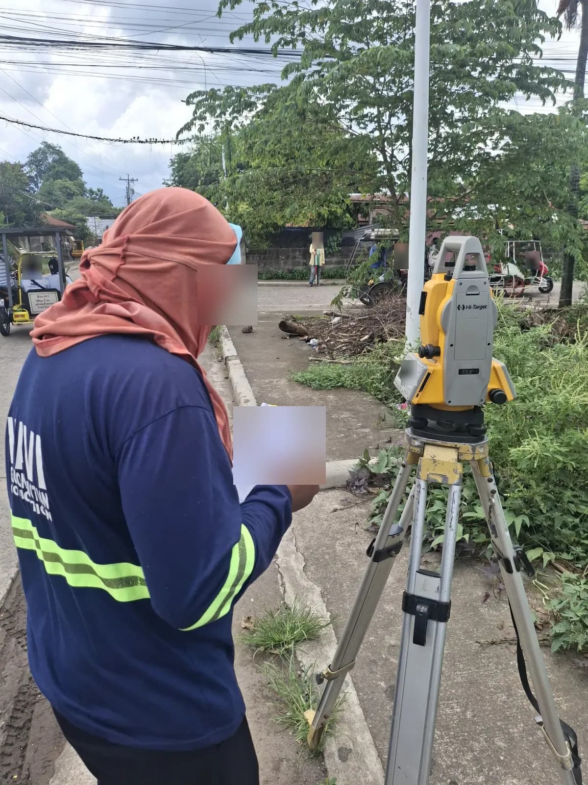 Survey operator writing in a notebook beside a total station with a prism-pole holder across the street