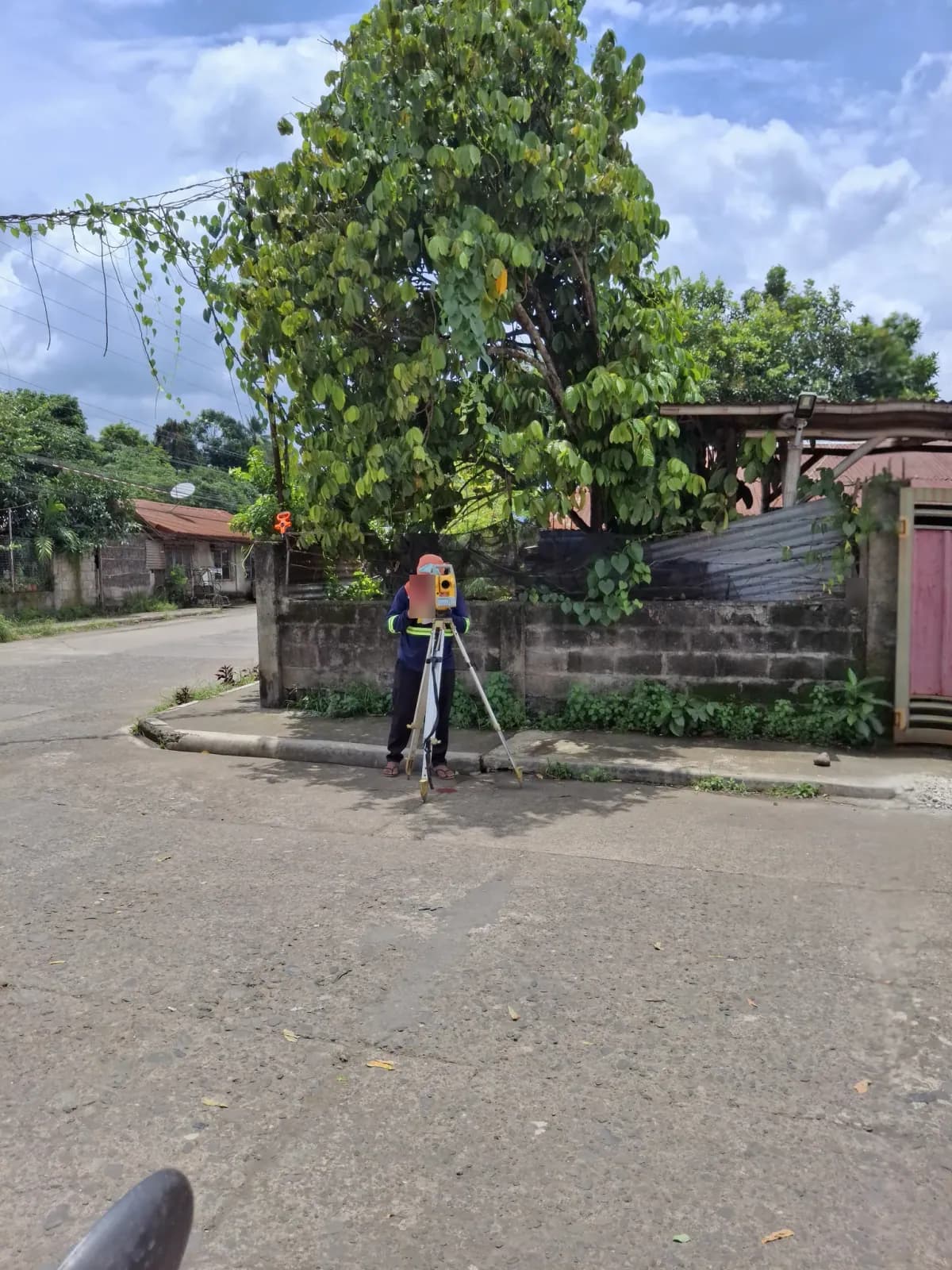 Wide view across the street toward a total-station operator working under a tree