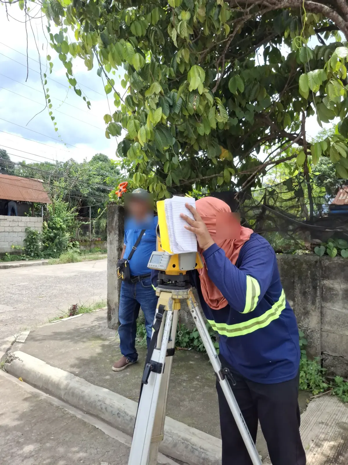 Survey operator sighting through a total station under a tree with a notebook beside the instrument