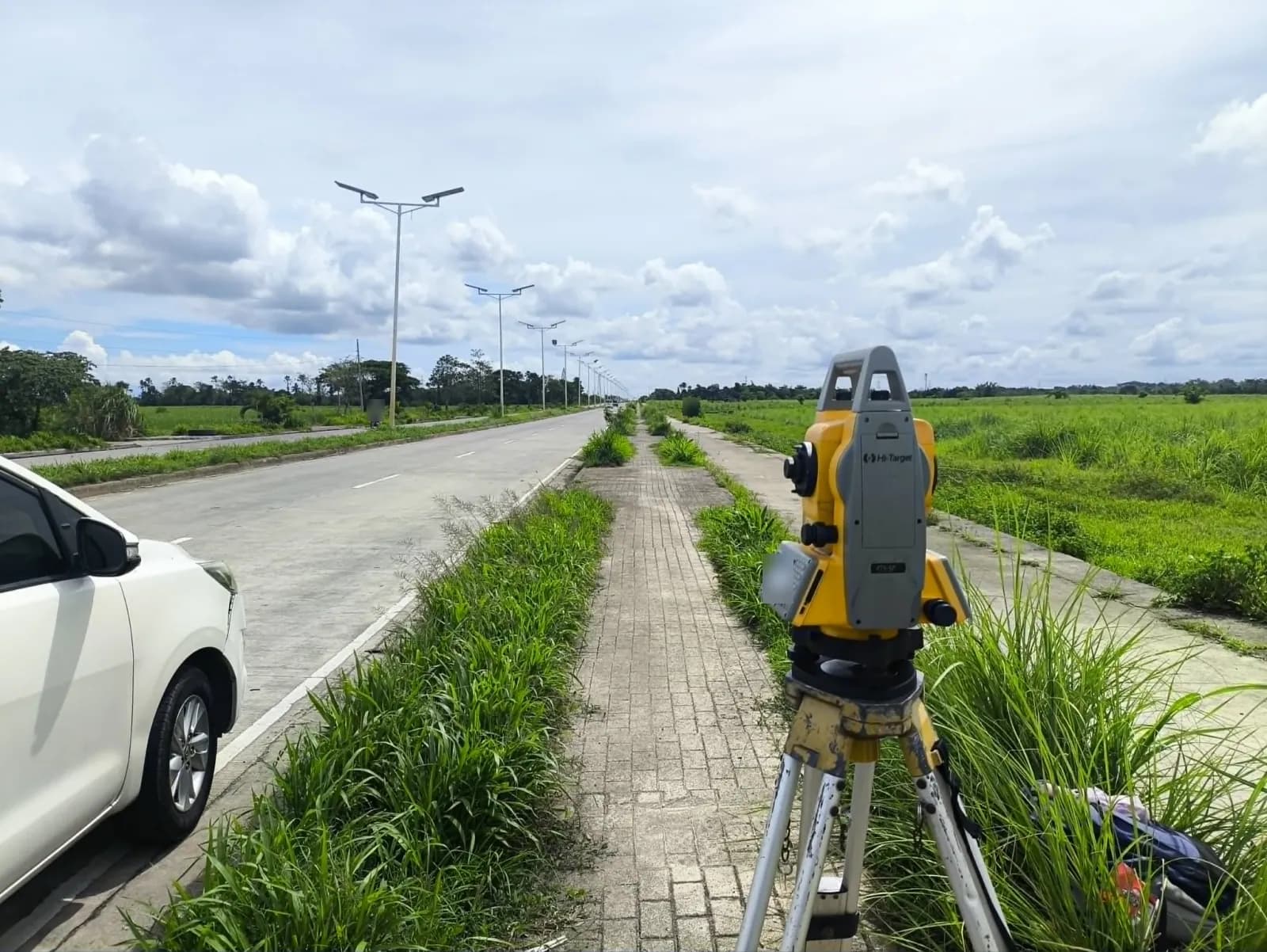 Total station on a sidewalk beside a divided road lined with streetlights, with open grassland and a parked white car nearby