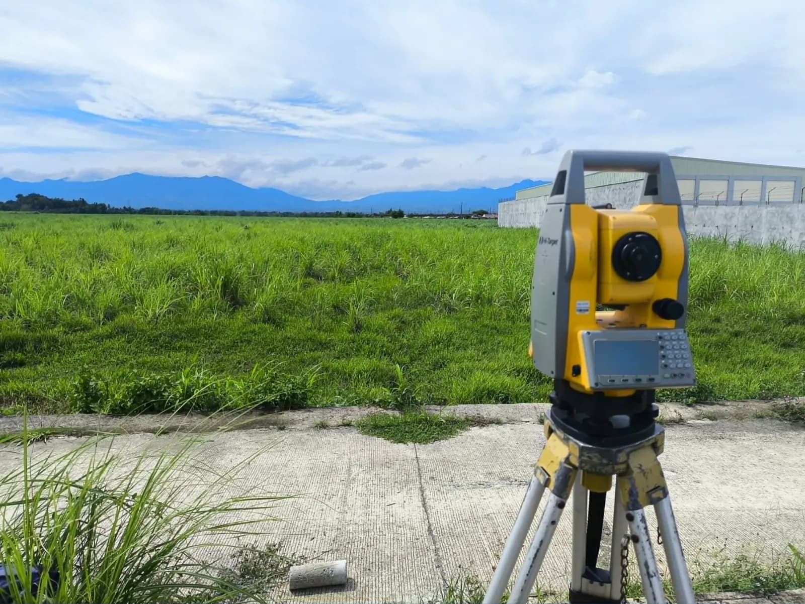 Total station on a tripod beside an open grassy lot in Bacolod, with a walled building and mountains in the distance