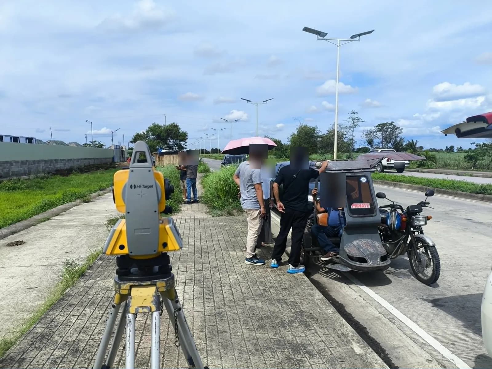 Total station in the foreground with people gathered beside parked vehicles and a tricycle along the road