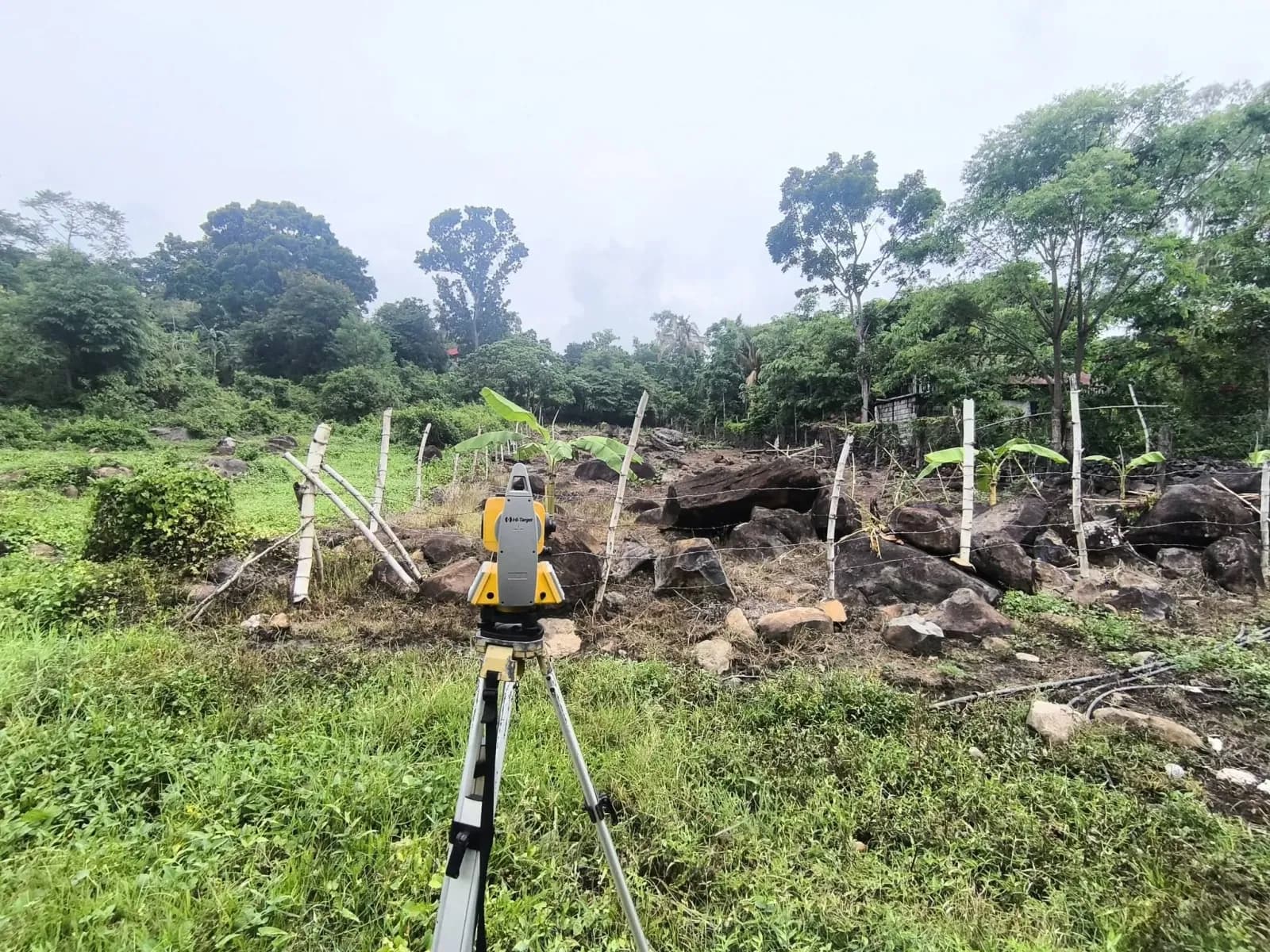 Tripod-mounted total station beside a fenced rocky site with large boulders, banana plants, and dense trees